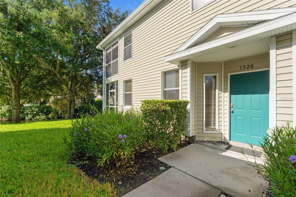 A two-story tan vinyl-sided building features a teal front door and a small landscaped garden — 1920 HAMMOCKS AVENUE UNIT 1002, LUTZ, FL