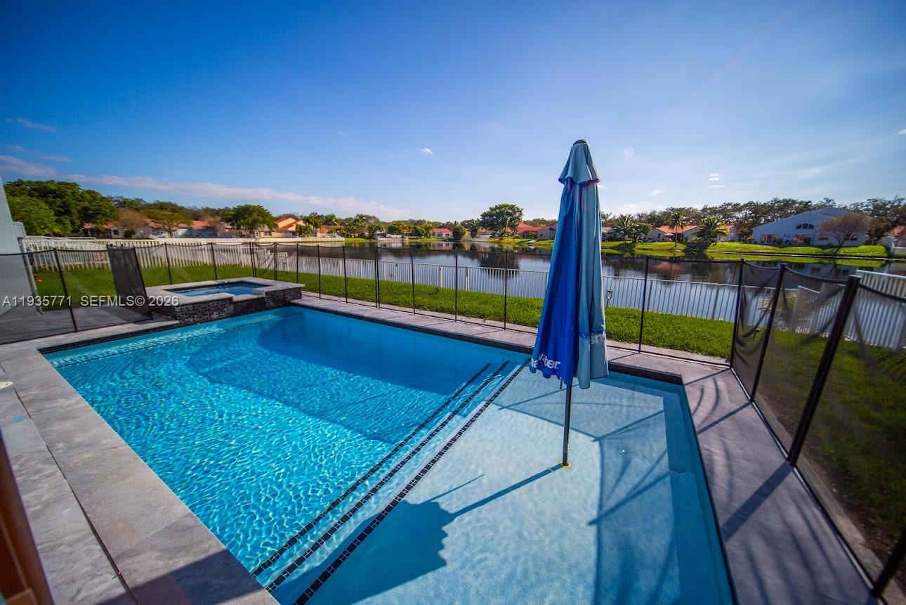 A swimming pool with a built-in spa and umbrella sits behind a fence overlooking a lake — 13020 SW 17th Ct, Miramar FL 33027, MIRAMAR, FL