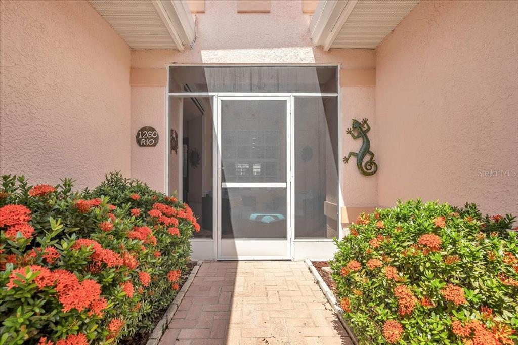 A paved walkway leads to a screened front entryway flanked by flowering bushes and stucco walls — 1260 RIO DE JANEIRO AVENUE UNIT 102, PUNTA GORDA, FL