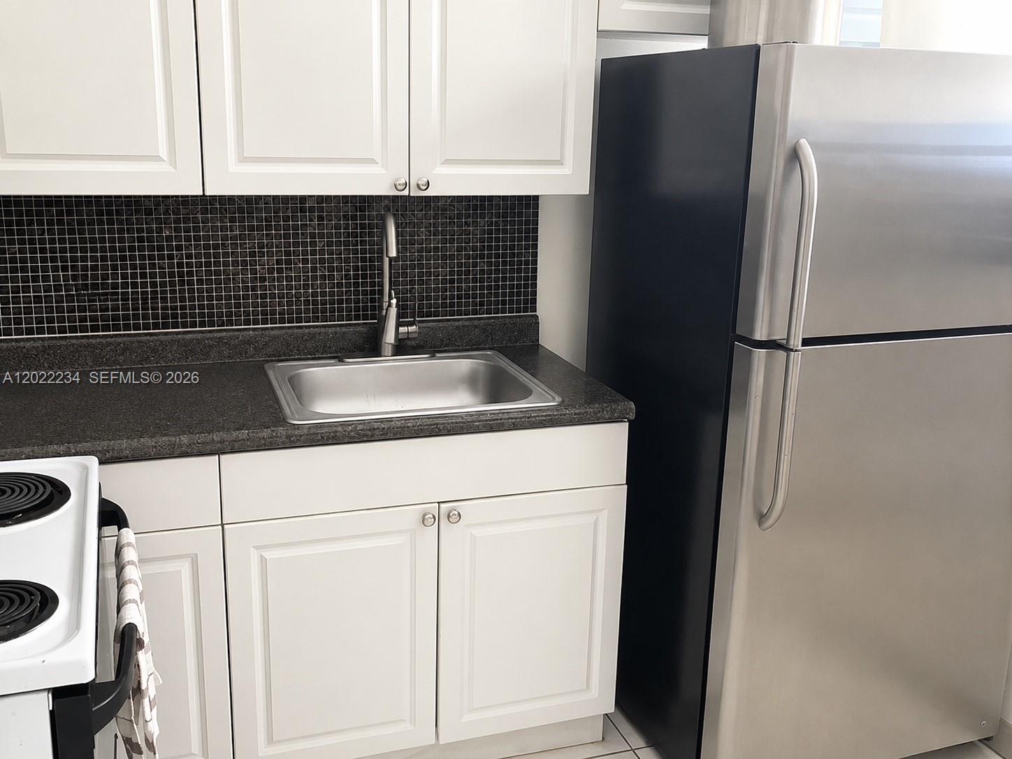 A kitchen features white cabinets, a stainless steel sink, dark countertops, and a stainless refrigerator — 716 NW 4th St, Hallandale Beach FL 33009, HALLANDALE BEACH, FL