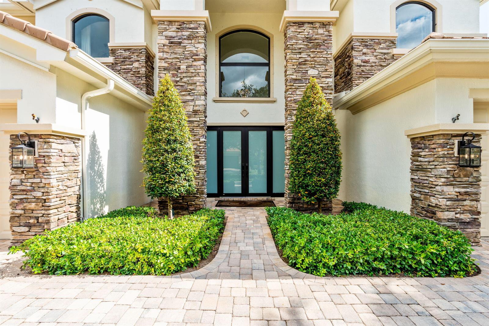 A stone-accented home entrance features a paved walkway, two conical trees, and double glass doors — 647 Edgebrook Lane, Royal Palm Beach FL 33411, ROYAL PALM BEACH, FL