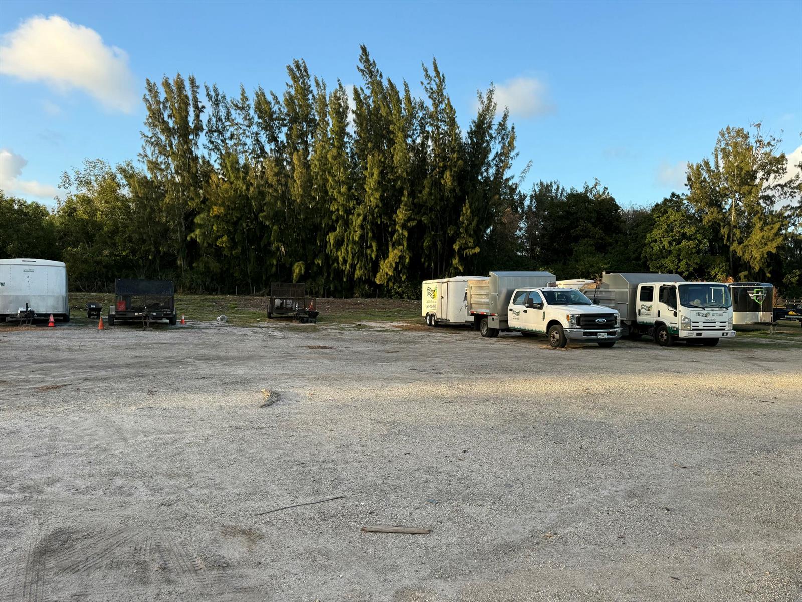 A gravel lot features several parked trailers and work trucks in front of a tree line — 9895 N 165th Street N, Jupiter FL 33478, JUPITER, FL
