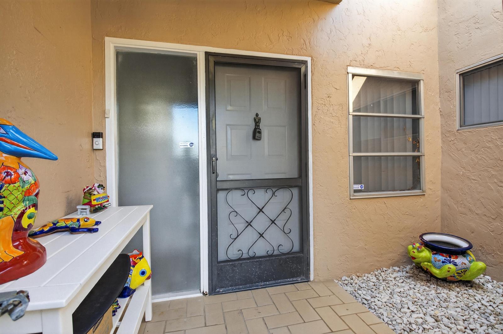 An exterior entryway features a white table with colorful decor, a screen door, and gravel — 8256 Summerbreeze Lane, Boca Raton FL 33496, BOCA RATON, FL