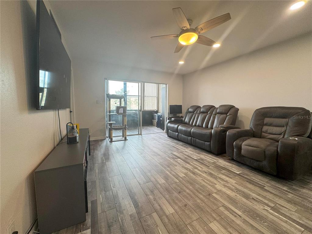 A living room features wood-look flooring, a ceiling fan, a large television, and leather furniture — 11485 OAKHURST ROAD UNIT -211, LARGO, FL