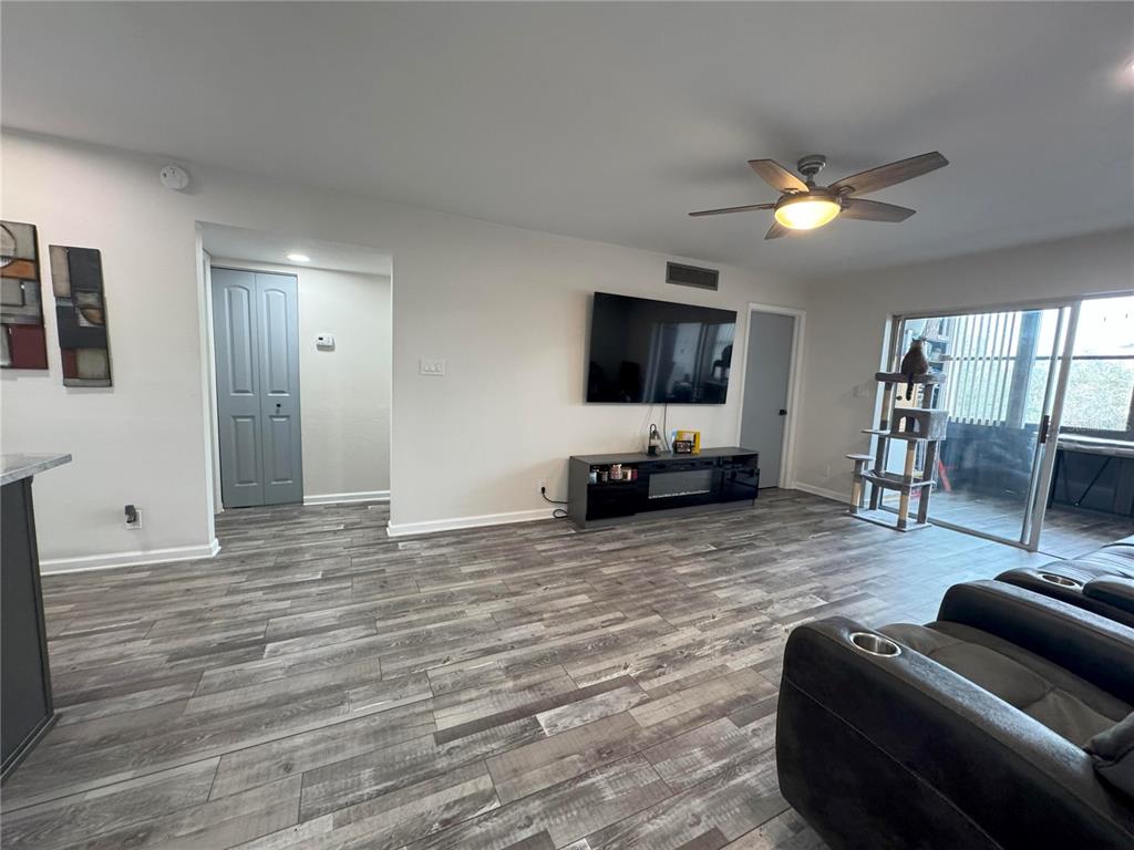 A living room features gray wood-look flooring, a wall-mounted television, and a sliding glass door — 11485 OAKHURST ROAD UNIT -211, LARGO, FL