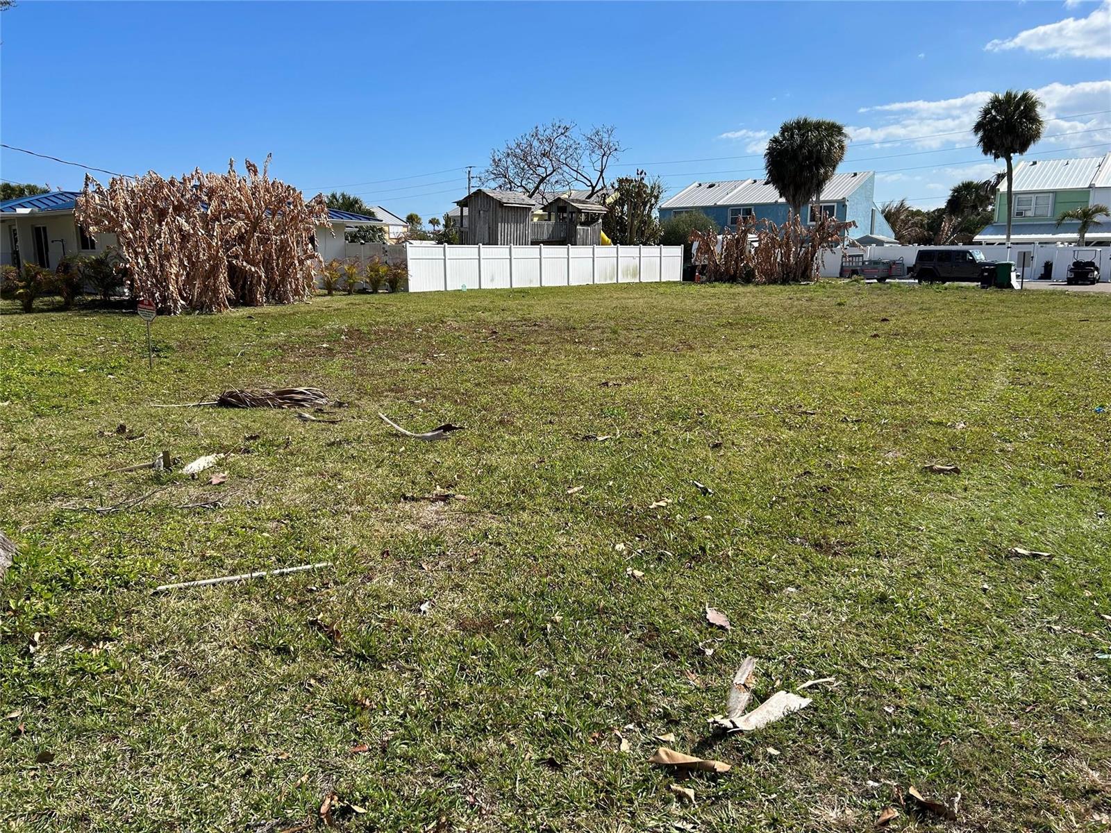 A vacant, grassy residential lot features a white fence and nearby houses under a blue sky — HUTCHINSON ISLAND, FL