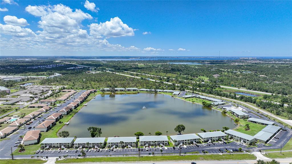 An aerial view shows a multi-building apartment complex surrounding a central lake with a fountain — 25225 RAMPART BOULEVARD UNIT 1607, PUNTA GORDA, FL