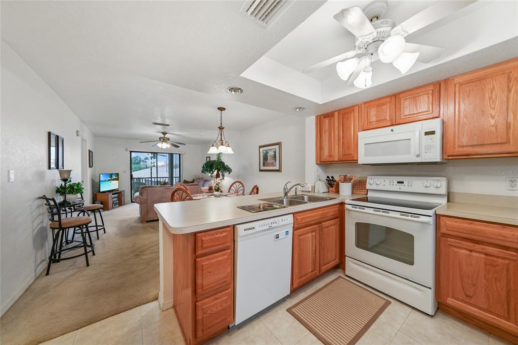 A kitchen with wood cabinets and white appliances opens into a carpeted living and dining area — 25225 RAMPART BOULEVARD UNIT 1607, PUNTA GORDA, FL