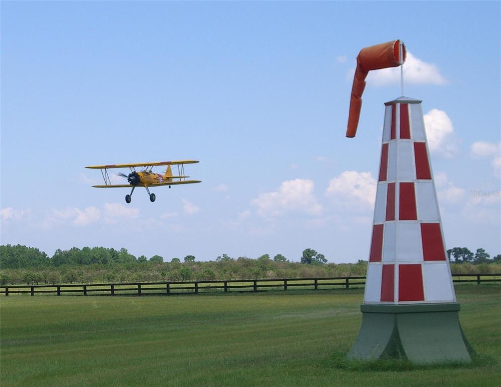 A yellow biplane flies over a grassy field near a red and white wind cone — GRASS ROOTS ROAD, GROVELAND, FL