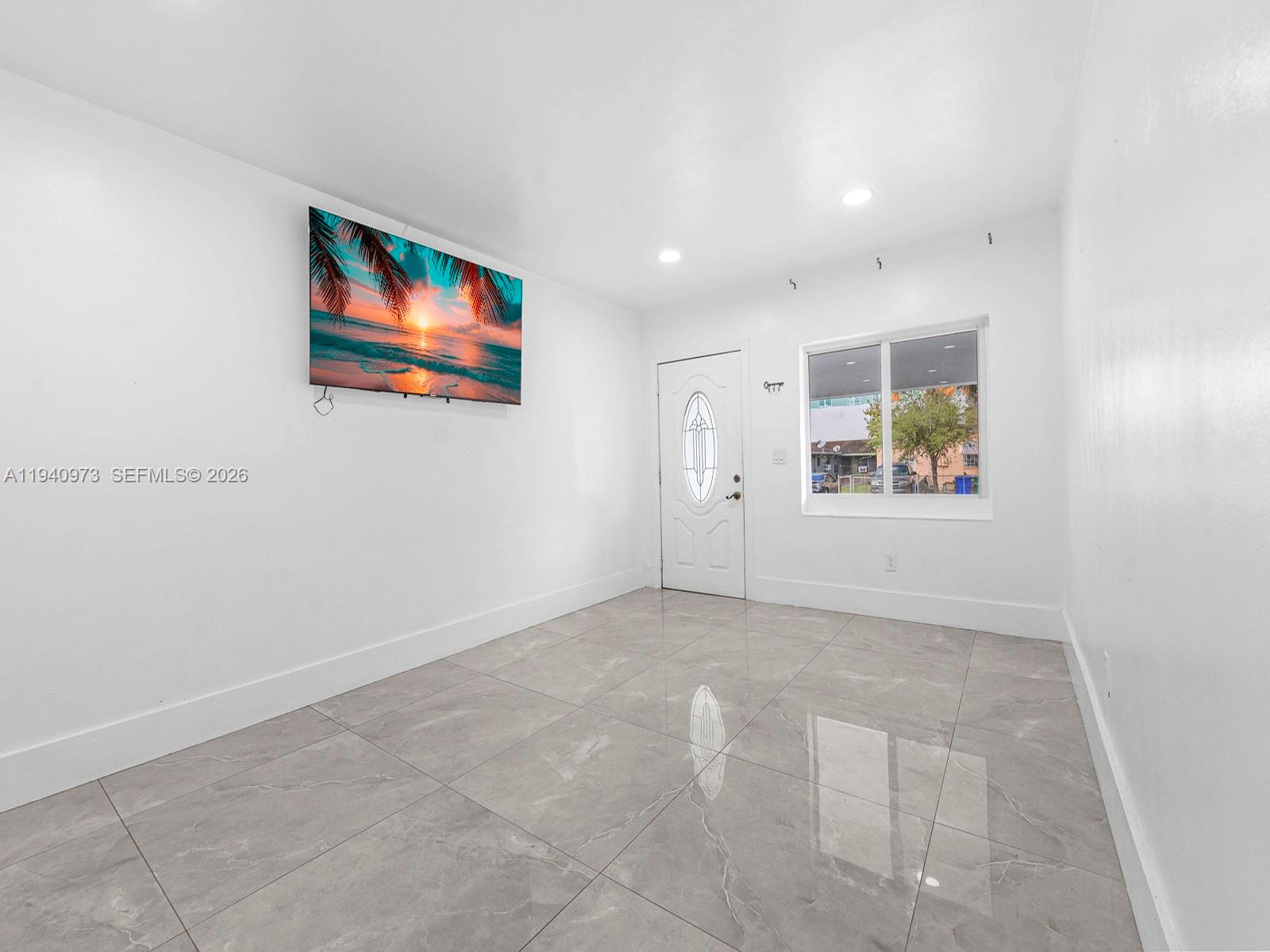 A bright room with white walls, gray tile flooring, a wall-mounted television, and an entry door — 1230 NW 35th St, Miami FL 33142, MIAMI, FL