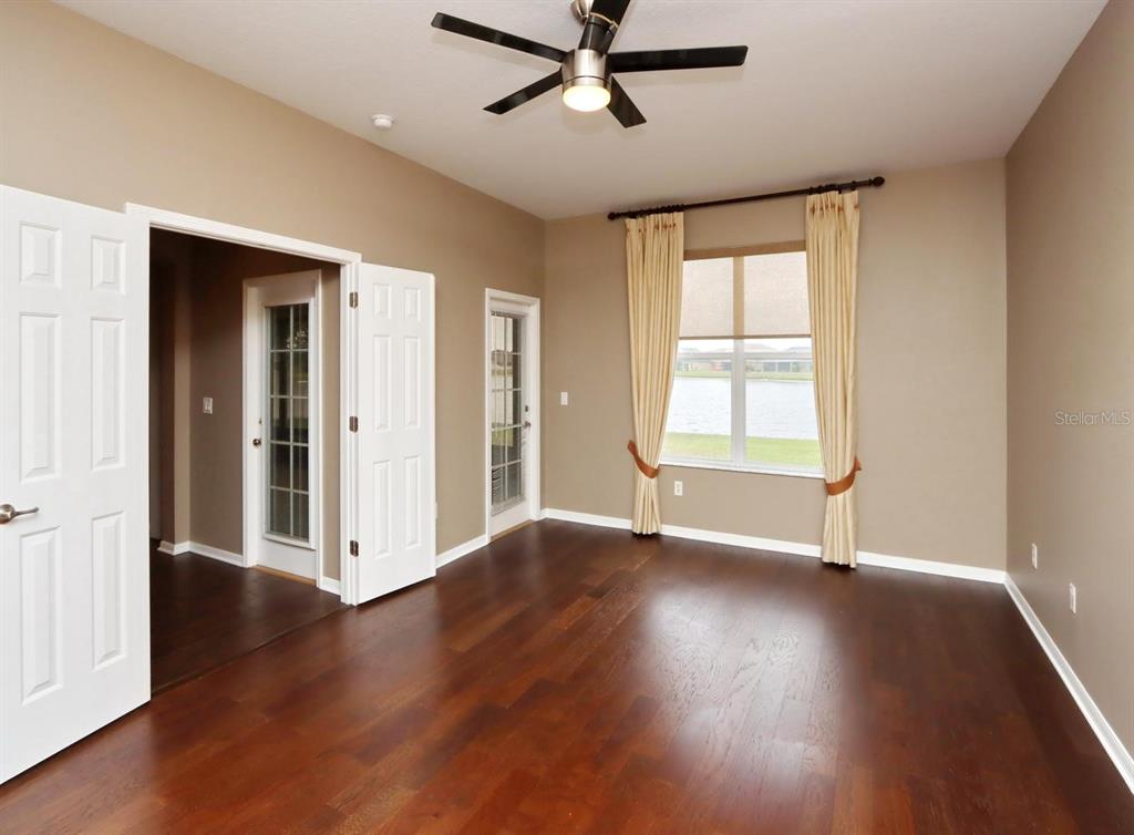 An empty room features dark wood flooring, tan walls, a ceiling fan, and a lake view — 13714 ARTESA BELL DRIVE, RIVERVIEW, FL
