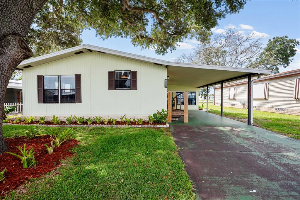 A light-colored mobile home features a carport, brown shutters, and a landscaped front yard — 36428 MALIBU WAY, ZEPHYRHILLS, FL