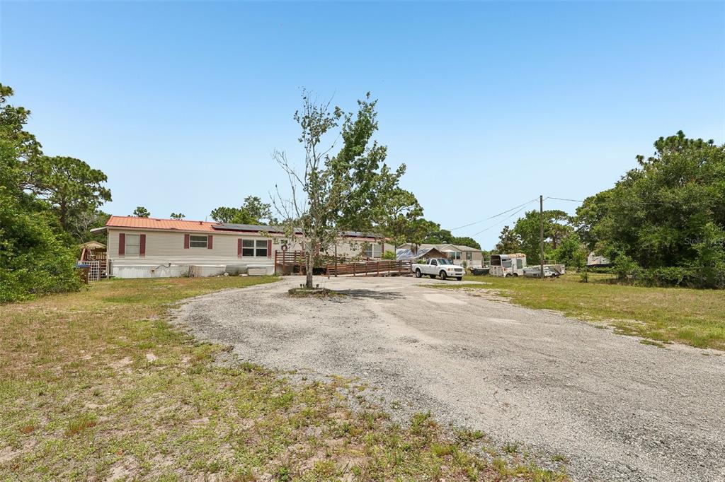 A single-story manufactured home sits on a large lot with a gravel driveway and trees — 9085 MERCURY DRIVE, ST CLOUD, FL