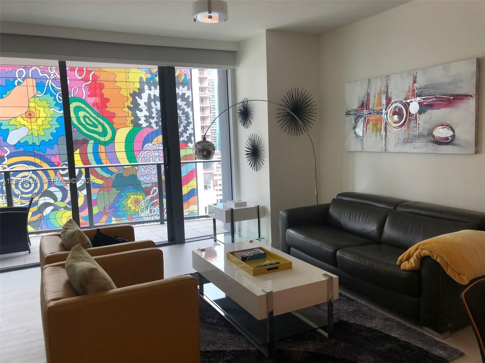 A living room features a dark leather sofa, two tan armchairs, and a vibrant mural outside — 1000 Brickell Plaza # 1914, Miami FL 33131, MIAMI, FL