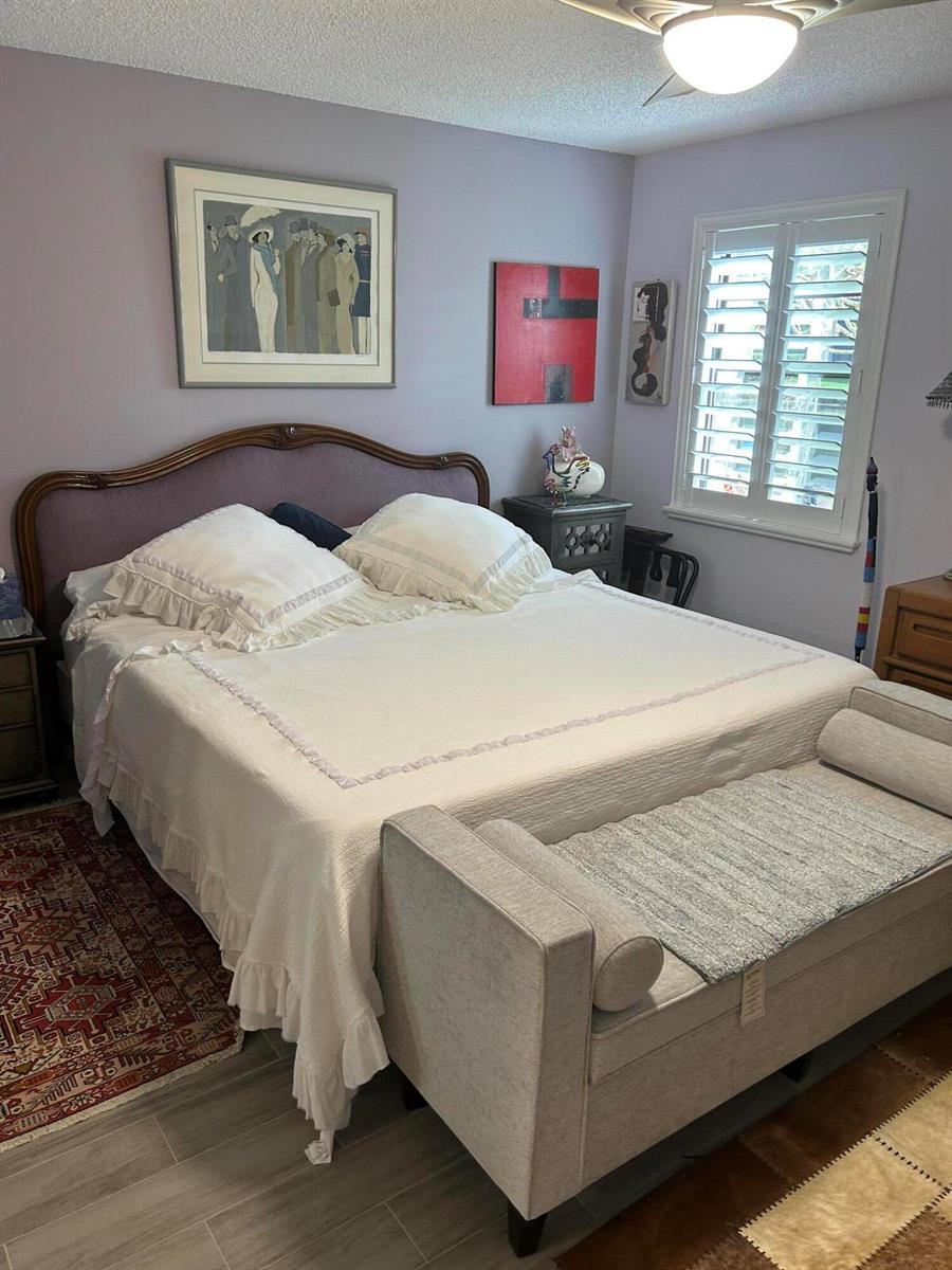 A bedroom features a bed with a ruffled white quilt, a grey bench, and wall art — 10271 N Circle Lake Drive, Boynton Beach FL 33437, BOYNTON BEACH, FL