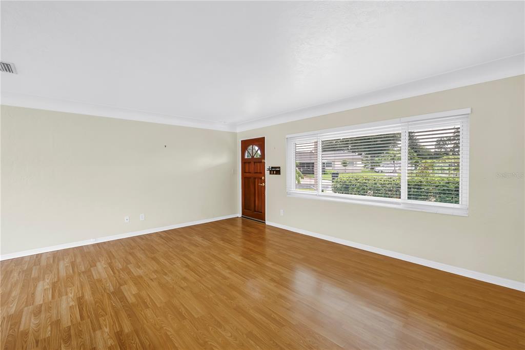 An empty living room features light-colored walls, wood-look flooring, a large window, and a door — 6195 28TH AVENUE N, ST PETERSBURG, FL