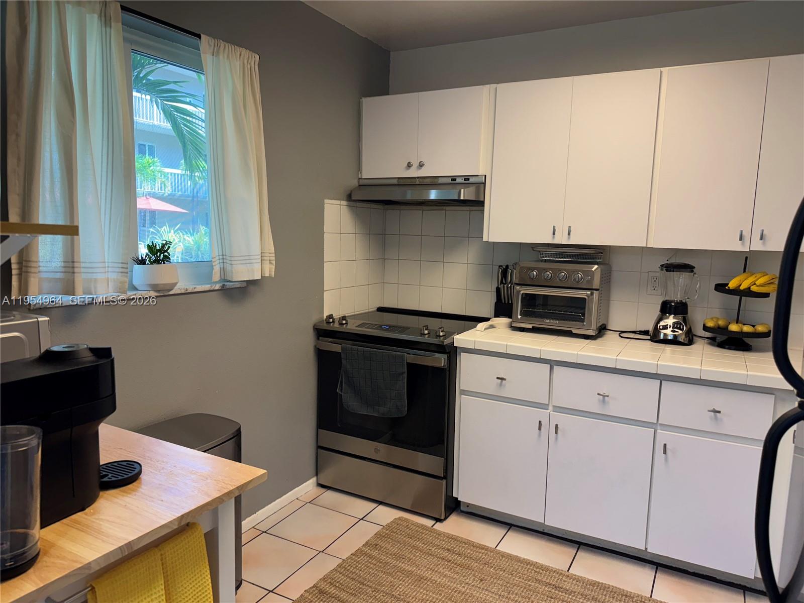 A kitchen features white cabinets, stainless steel appliances, tiled countertops, and a window with curtains — 7703 Camino Real # A106, Miami FL 33143, MIAMI, FL
