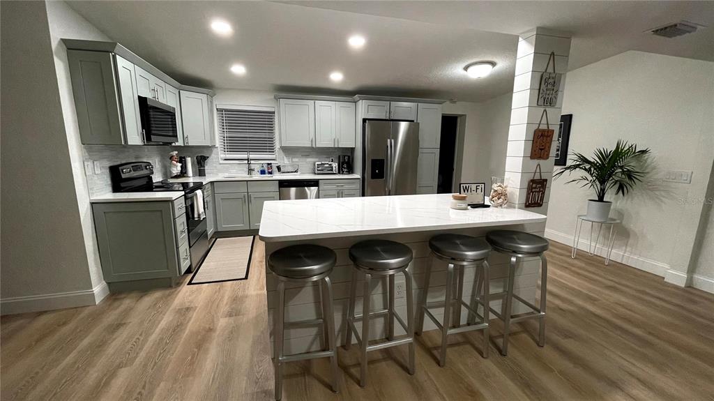 A kitchen features light gray cabinets, a white quartz island with stools, and wood flooring — 1639 ST CATHERINE DRIVE W, DUNEDIN, FL