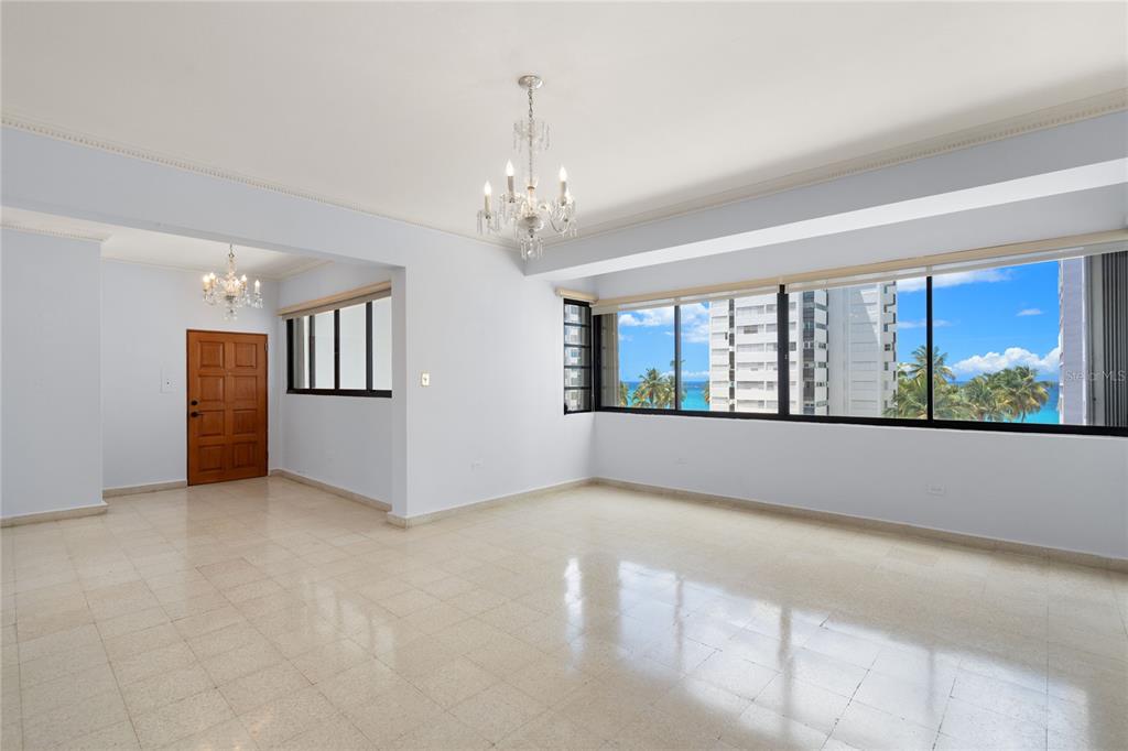 An empty living room with light tile floors, two chandeliers, and large windows overlooking the ocean — 61 KINGS COURT UNIT 5E, SAN JUAN, PR