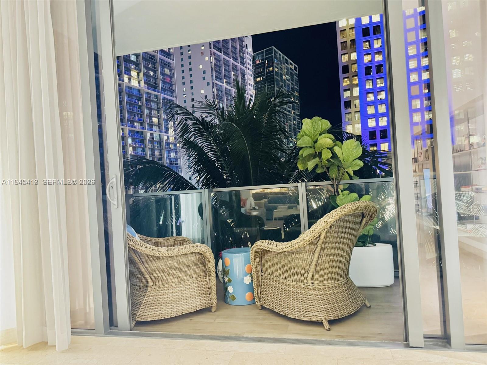 A balcony with two wicker chairs, a small blue table, and a potted plant at night — 68 SE 6th St # 903, Miami FL 33131, MIAMI, FL