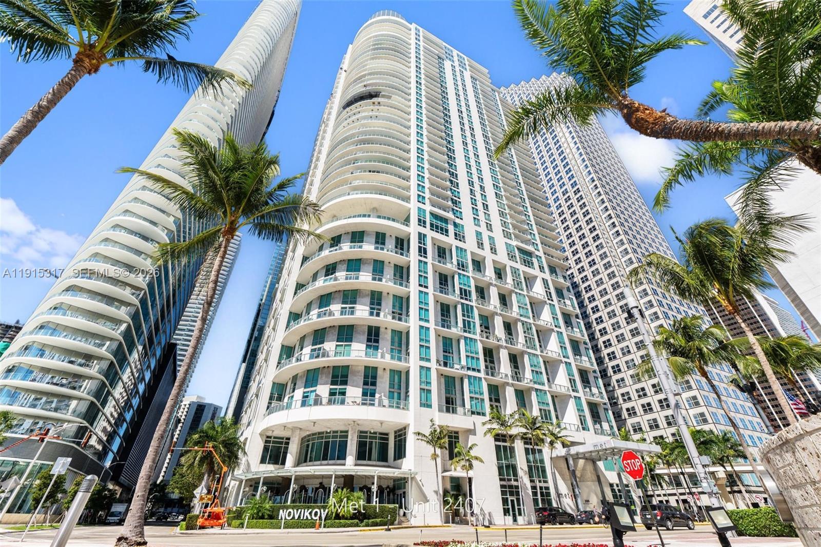 A low-angle exterior view shows a modern high-rise residential building with curved balconies and palm trees — 300 S Biscayne Blvd # 2407, Miami FL 33131, MIAMI, FL