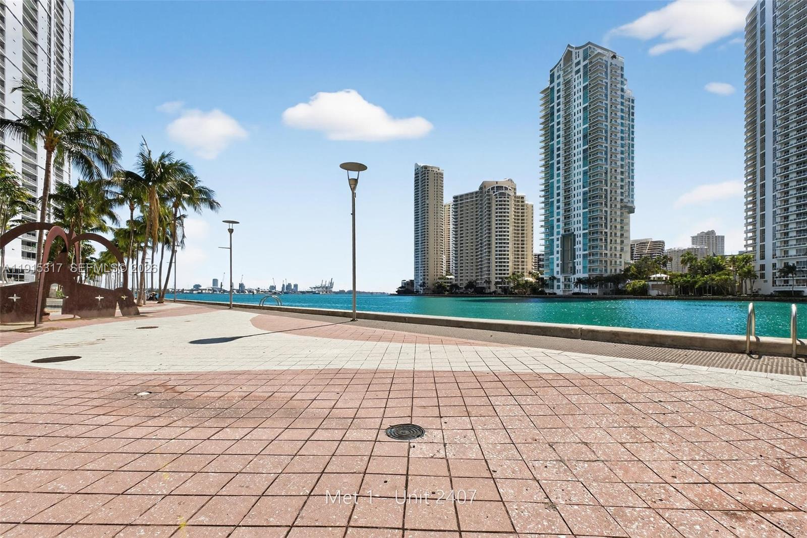 A paved waterfront promenade features palm trees, modern high-rise buildings, and a view of the bay — 300 S Biscayne Blvd # 2407, Miami FL 33131, MIAMI, FL