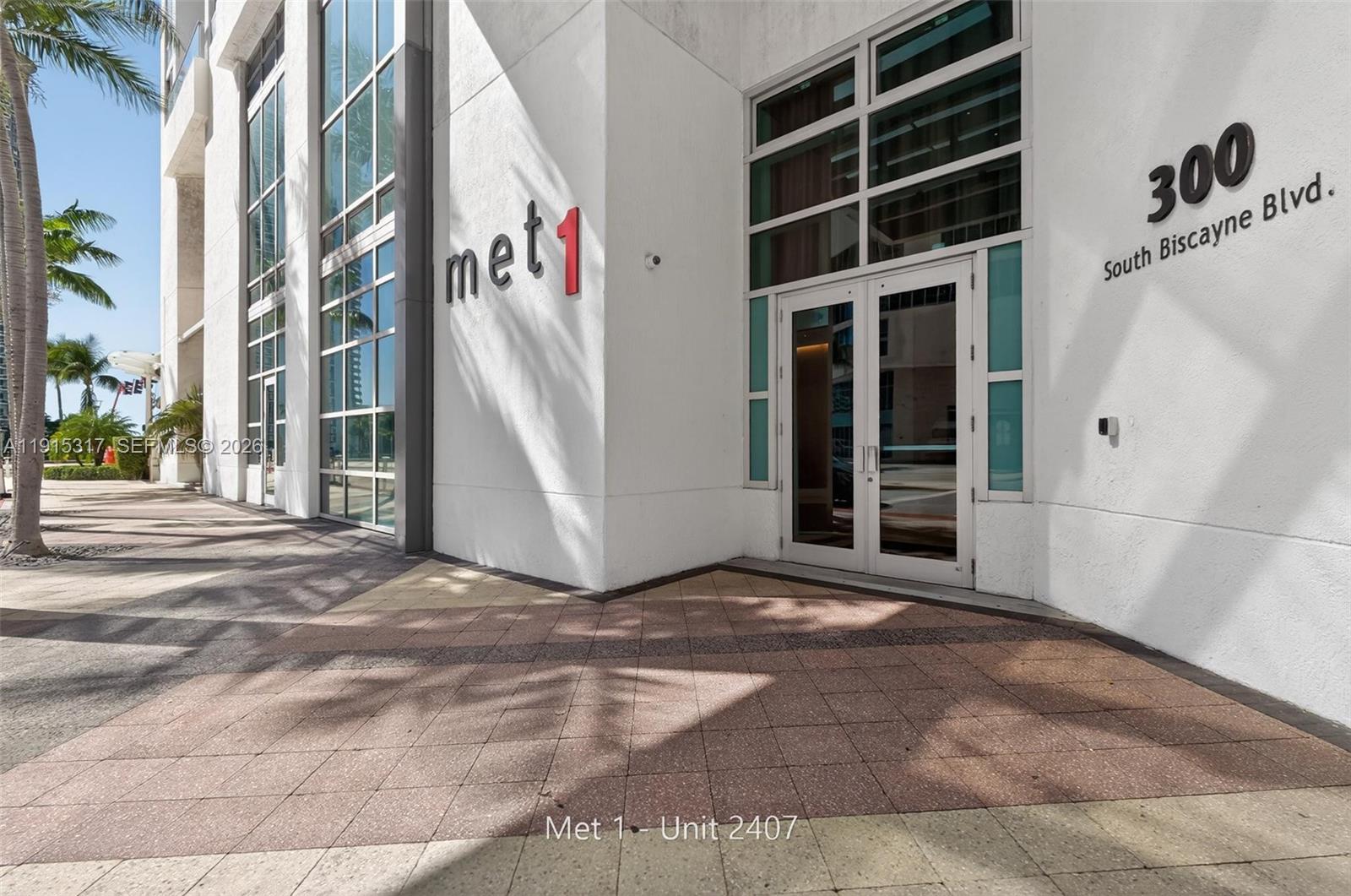 The exterior entrance of a modern building features glass doors and a tiled walkway — 300 S Biscayne Blvd # 2407, Miami FL 33131, MIAMI, FL