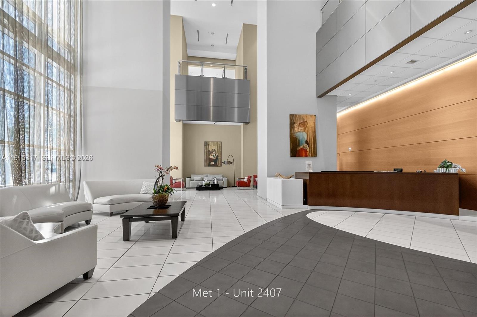 A modern building lobby features high ceilings, white tile floors, and a dark wood reception desk — 300 S Biscayne Blvd # 2407, Miami FL 33131, MIAMI, FL