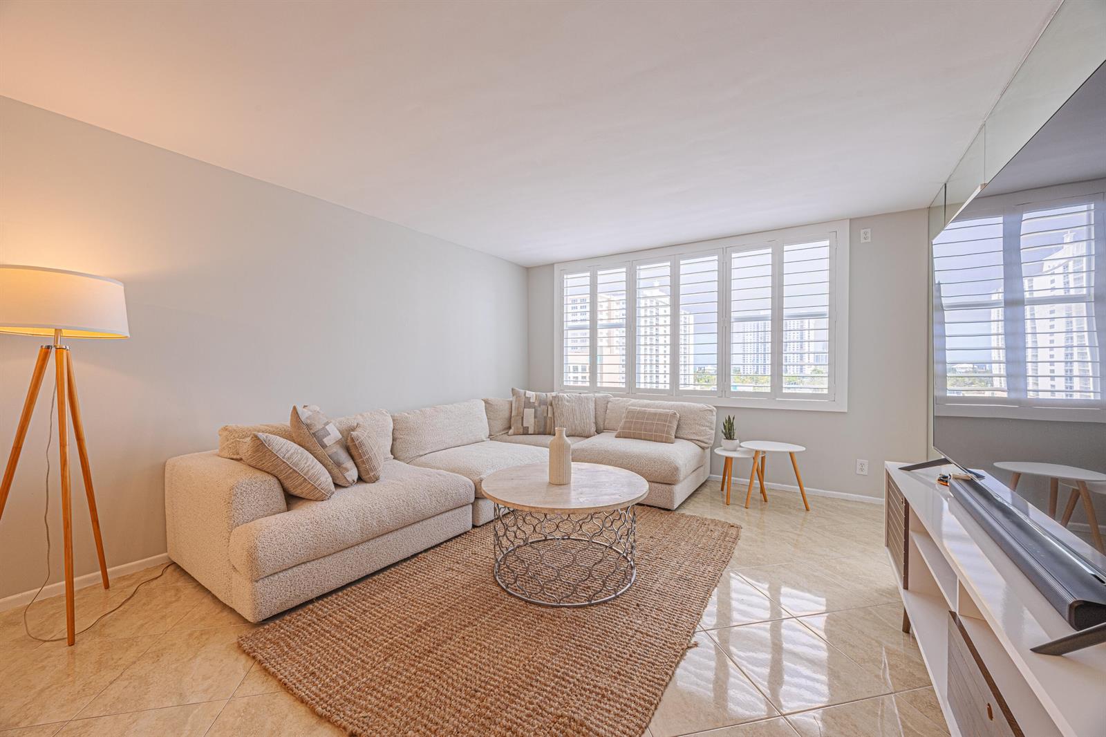A living room features a beige sectional sofa, a round coffee table, and plantation shutters — 201 N Ocean Boulevard # 702, Pompano Beach FL 33062, POMPANO BEACH, FL