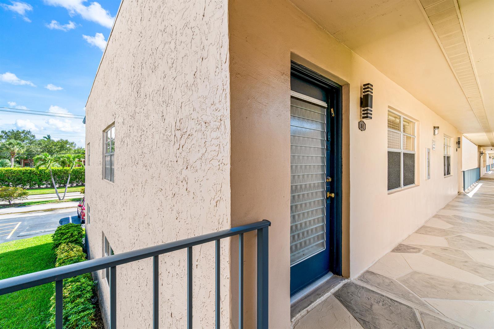 An exterior view of an apartment building walkway featuring a dark blue door and stucco walls — 184 Flanders D, Delray Beach FL 33484, DELRAY BEACH, FL