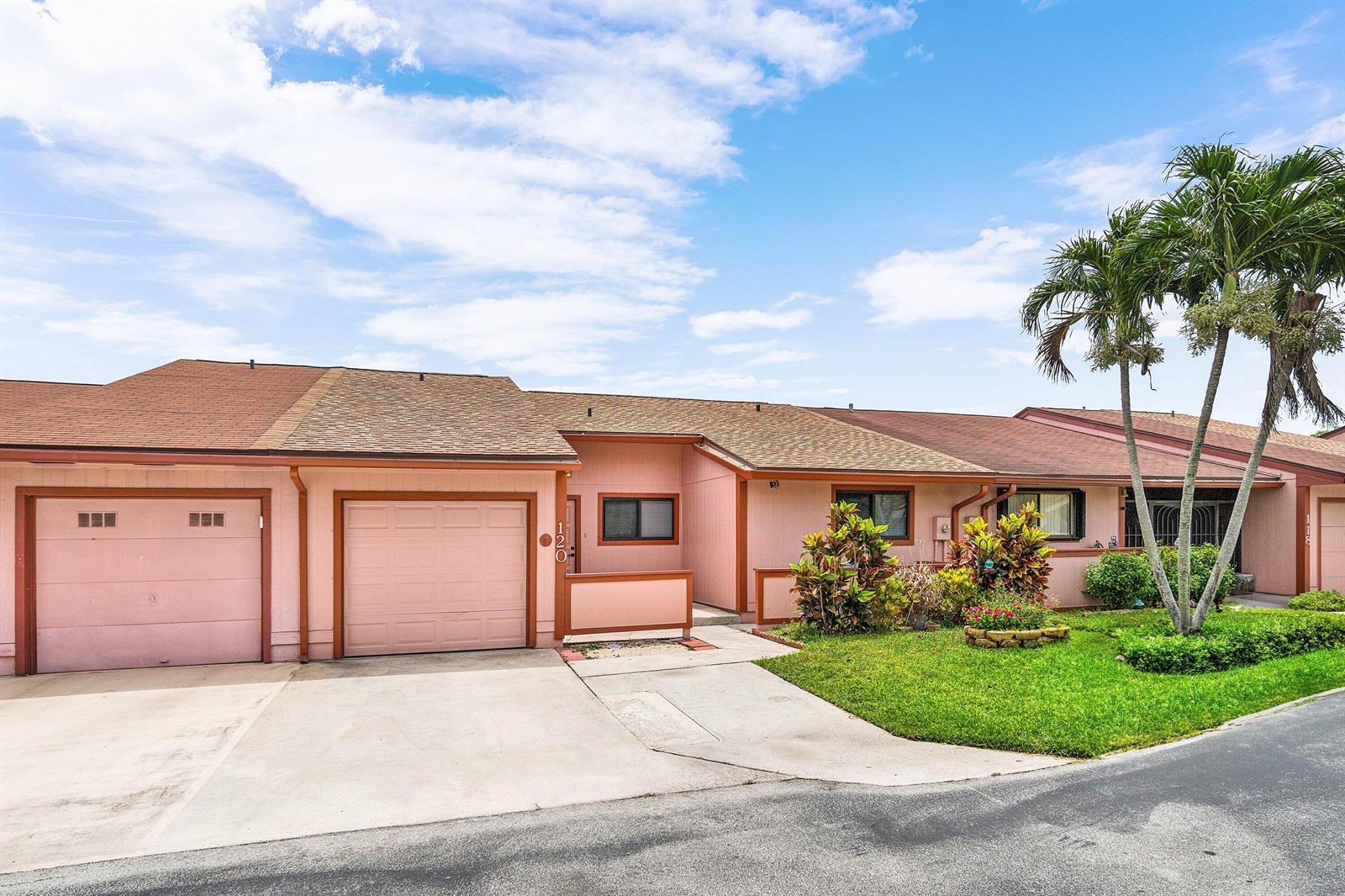 A pink, single-story attached house features a two-car garage, a concrete driveway, and palm trees — 120 Mayfair Lane, Boynton Beach FL 33426, BOYNTON BEACH, FL
