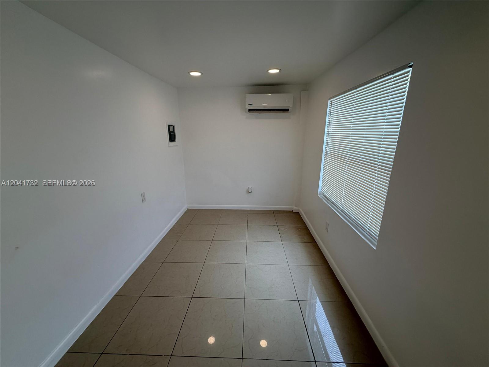 A small, empty room features white walls, tan tiled flooring, a window with blinds, and a wall-mounted air conditioner — 548 NW 30th St # 2, Miami FL 33127, MIAMI, FL