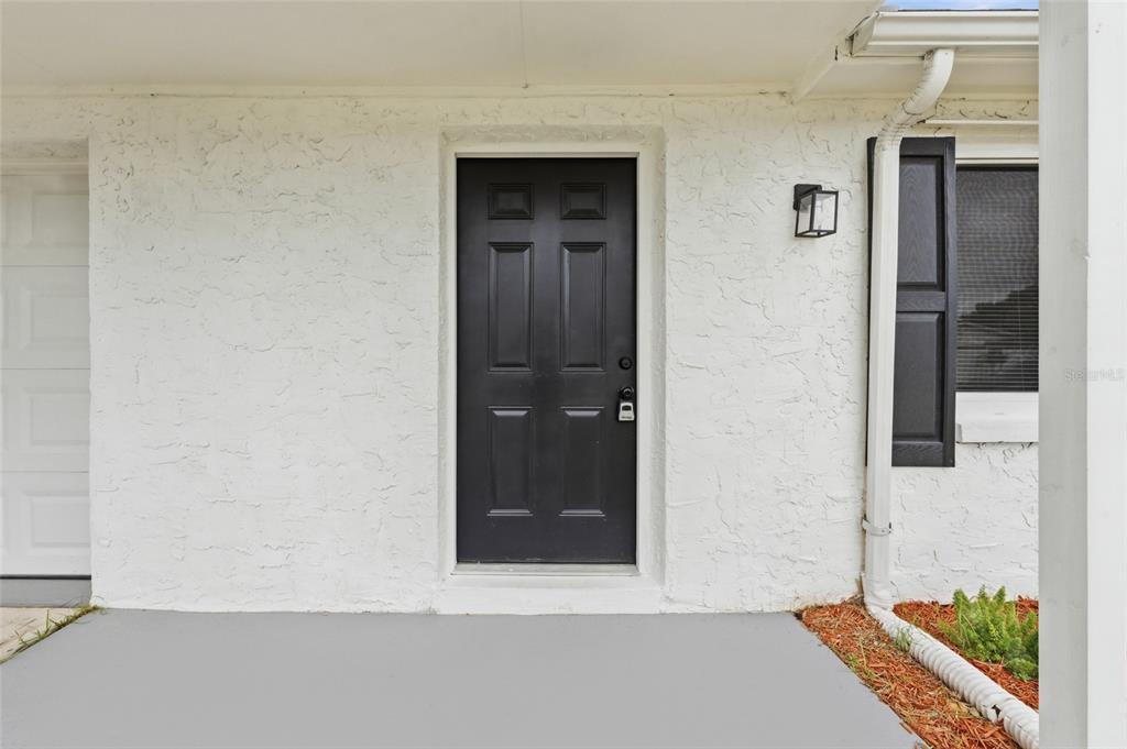 A black front door is centered on a white stucco wall next to a window — 3306 CANTRELL STREET, HOLIDAY, FL