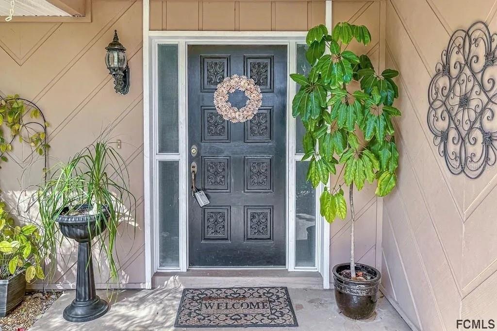 A dark front door with a wreath and sidelights is flanked by potted plants — 13 CEDARFORD COURT, PALM COAST, FL