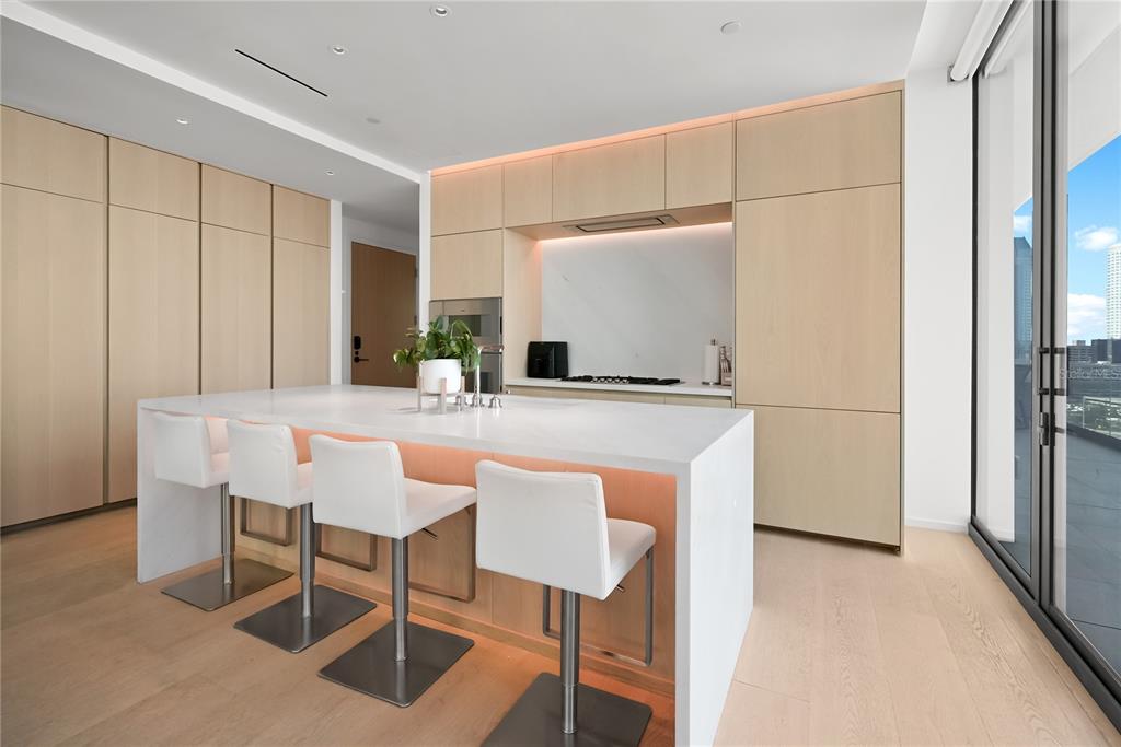 A modern kitchen features light wood cabinetry, a white waterfall island, and white bar stools — 1000 WATER STREET UNIT 1002, TAMPA, FL