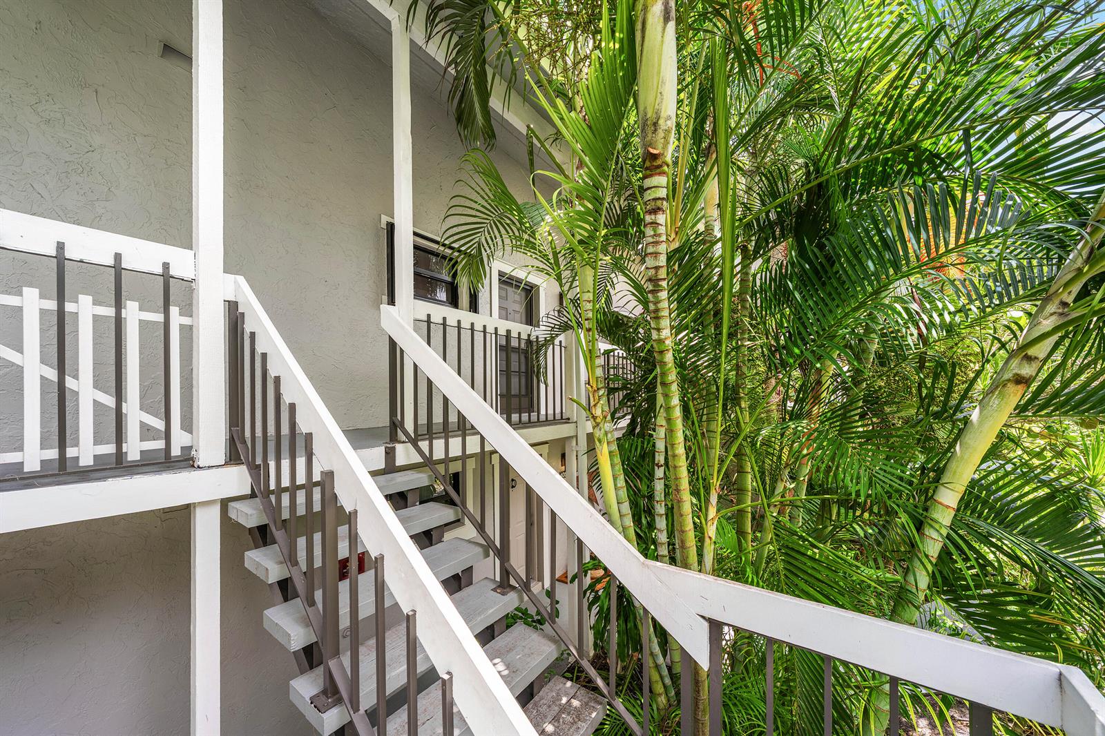 An outdoor staircase with white railings leads up to a building entrance beside palm trees — 11863 Wimbledon Circle # 504, Wellington FL 33414, WELLINGTON, FL