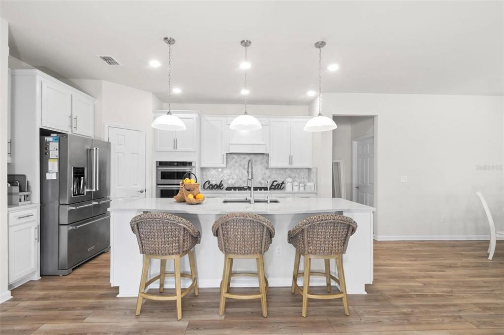 A modern kitchen features white cabinetry, a large island with three wicker stools, and stainless appliances — 12457 GOLDEN SAGE DRIVE, SARASOTA, FL