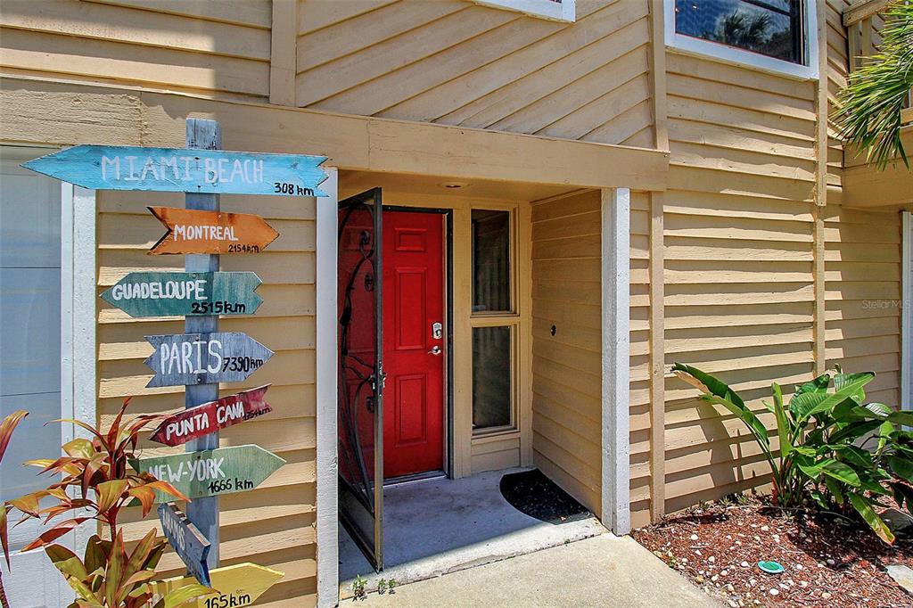 A red front door is adjacent to a wooden post displaying directional signs with distances — 4003 BAYSIDE COURT, BRADENTON, FL