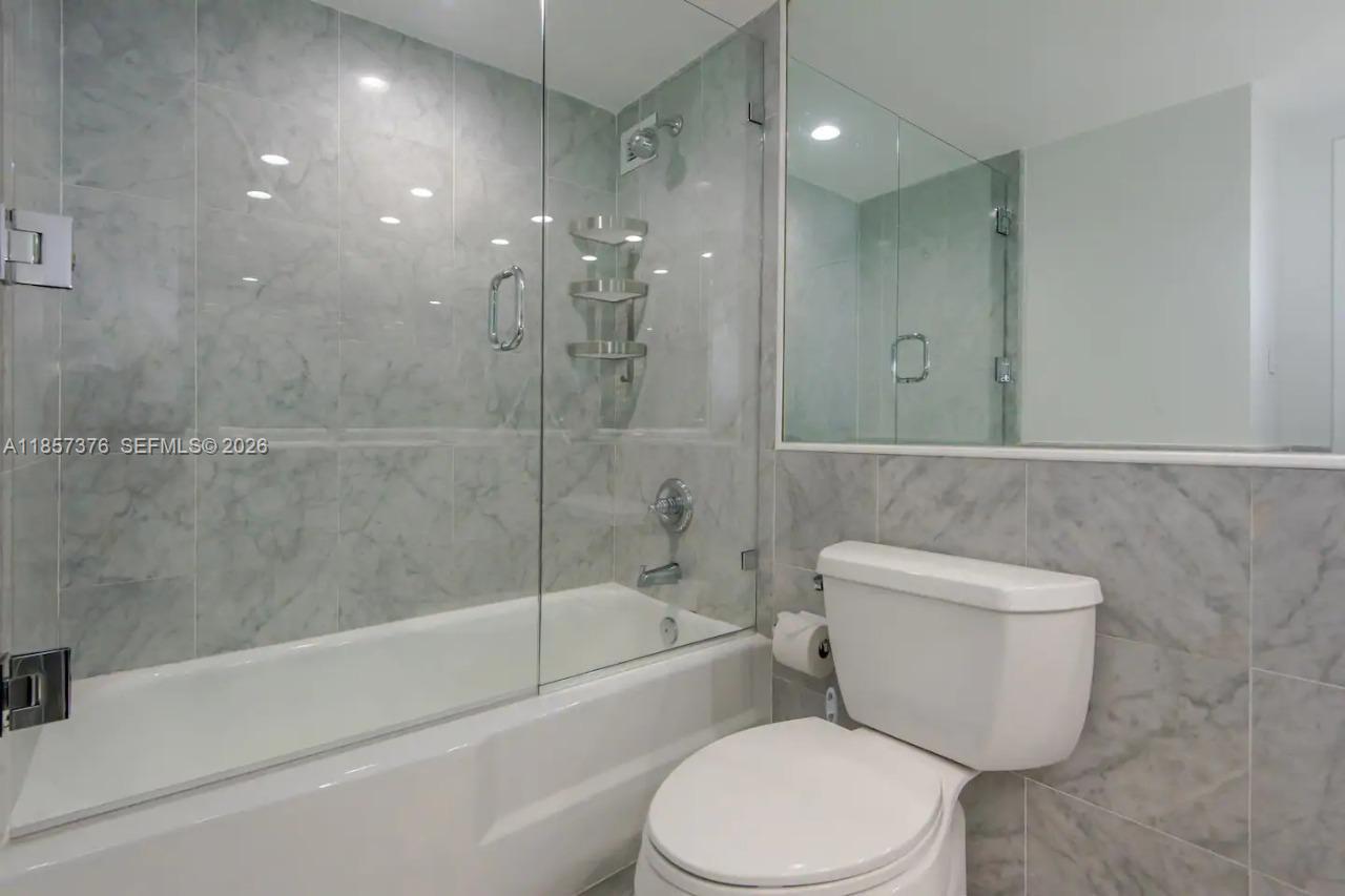 A bathroom features a white toilet and a bathtub with a glass enclosure and marble tiles — 5161 Collins Ave # 1104, Miami Beach FL 33140, MIAMI BEACH, FL