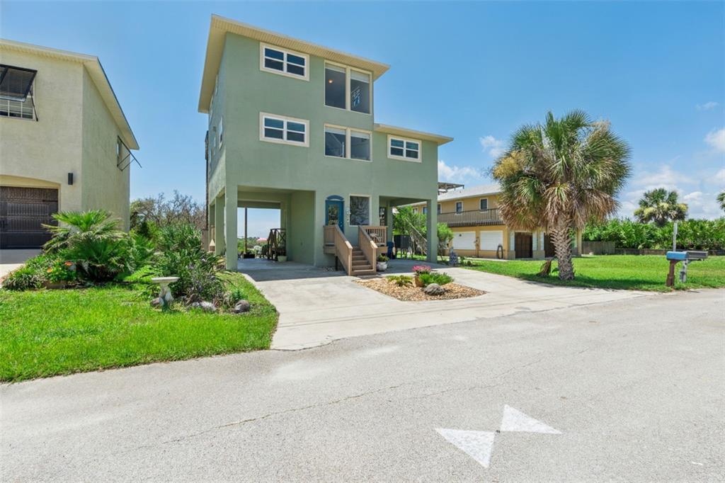 A three-story green stilt house with a concrete driveway and a palm tree in front — 884 CATFISH AVENUE, NEW SMYRNA BEACH, FL