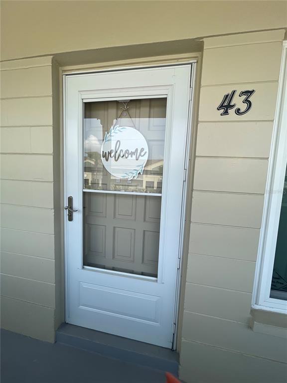 A white storm door with a welcome sign is mounted on a beige exterior wall — 2295 BELGIAN LANE UNIT 43, CLEARWATER, FL