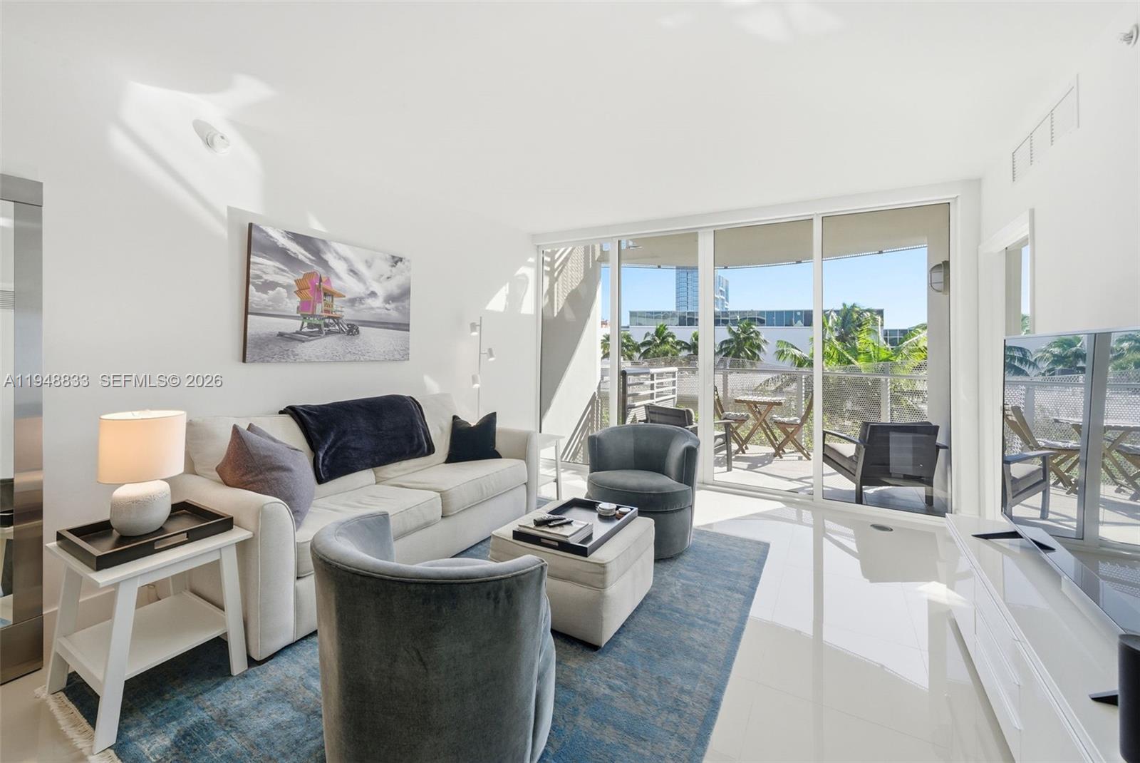 A bright living room features a white sofa, blue armchairs, and a balcony with outdoor seating — 2155 Washington Ct # 303, Miami Beach FL 33139, MIAMI BEACH, FL