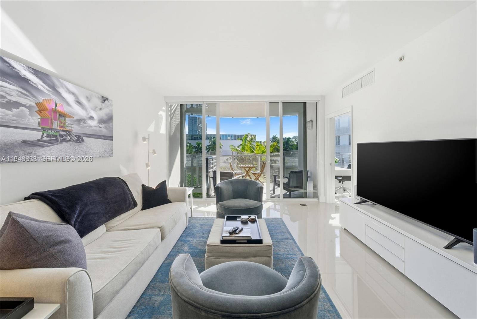 A modern living room features a white sofa, two gray armchairs, and a large television — 2155 Washington Ct # 303, Miami Beach FL 33139, MIAMI BEACH, FL