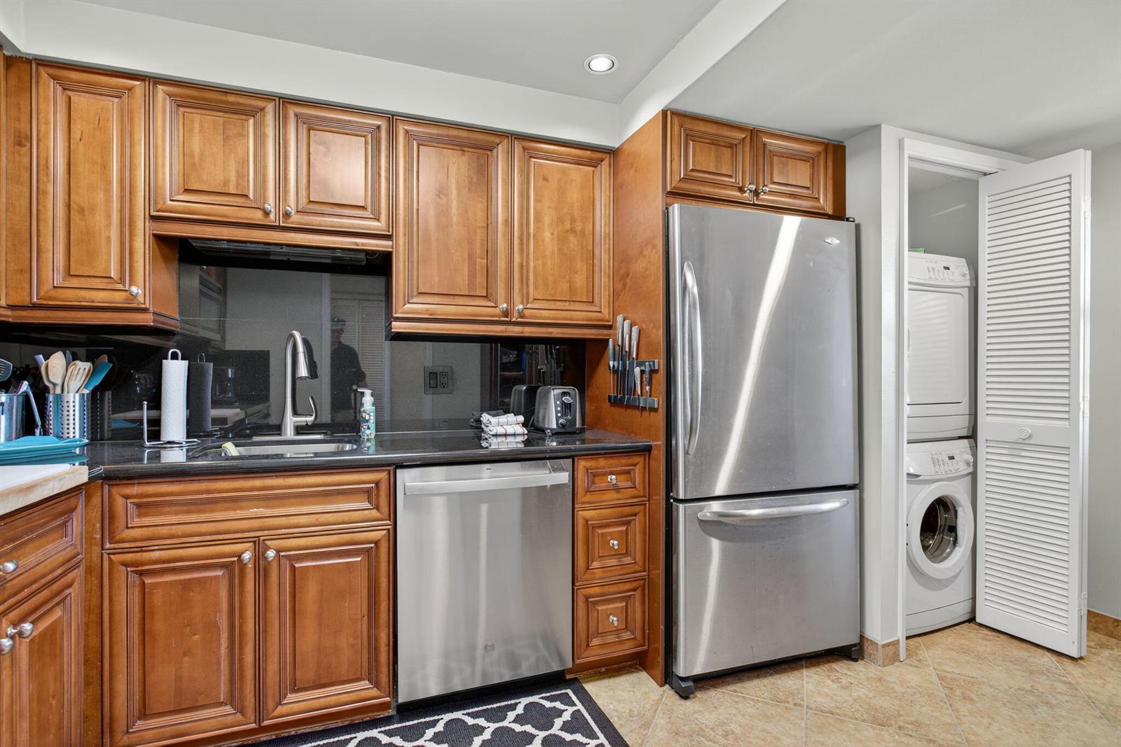 A kitchen features wood cabinets, stainless steel appliances, black countertops, and an adjacent laundry closet — 2900 N Course Drive # 905, Pompano Beach FL 33069, POMPANO BEACH, FL