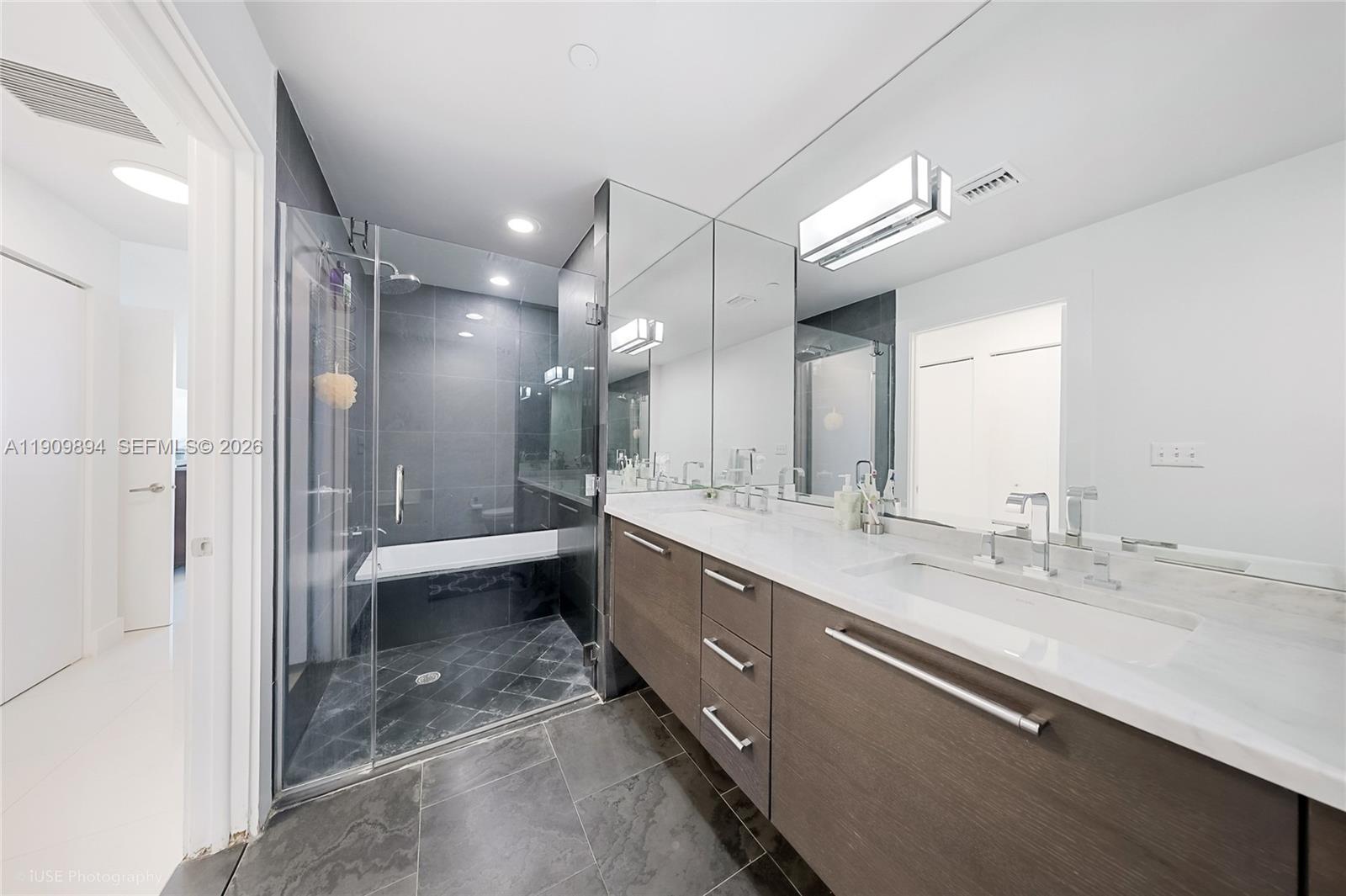A modern bathroom features a glass-enclosed shower-tub combination, a double vanity, and dark tile flooring — 900 Biscayne Blvd # 2602, Miami FL 33132, MIAMI, FL