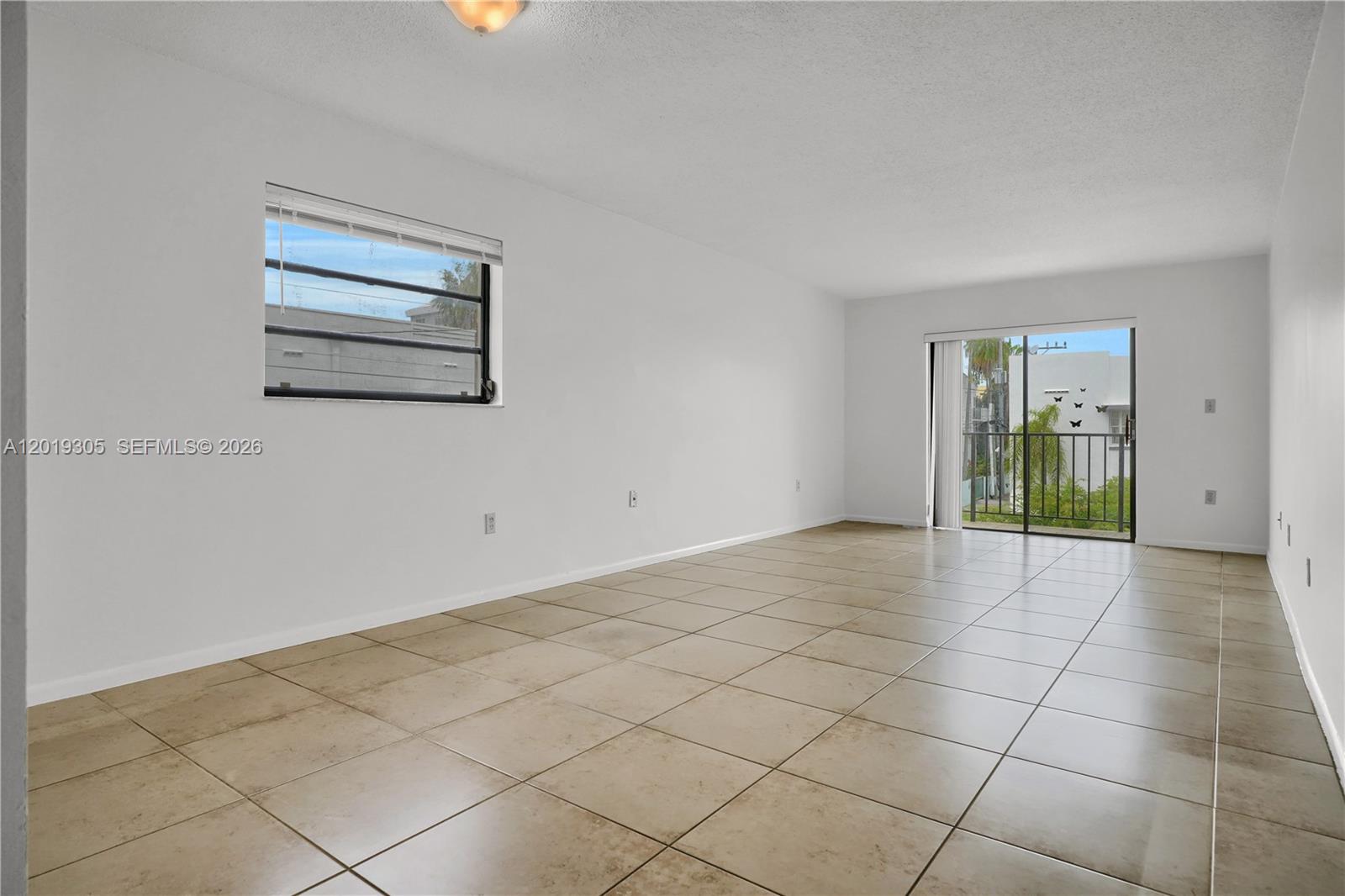 An empty living room features white walls, beige tile flooring, a window, and a balcony door — 950 Euclid Ave # 201, Miami Beach FL 33139, MIAMI BEACH, FL