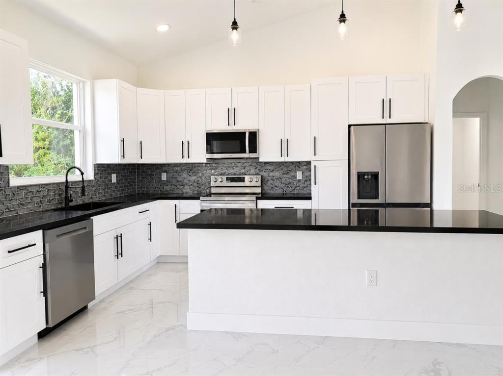 A modern kitchen features white cabinets, black countertops, a gray tile backsplash, and stainless appliances — 3476 SE HWY 484, BELLEVIEW, FL