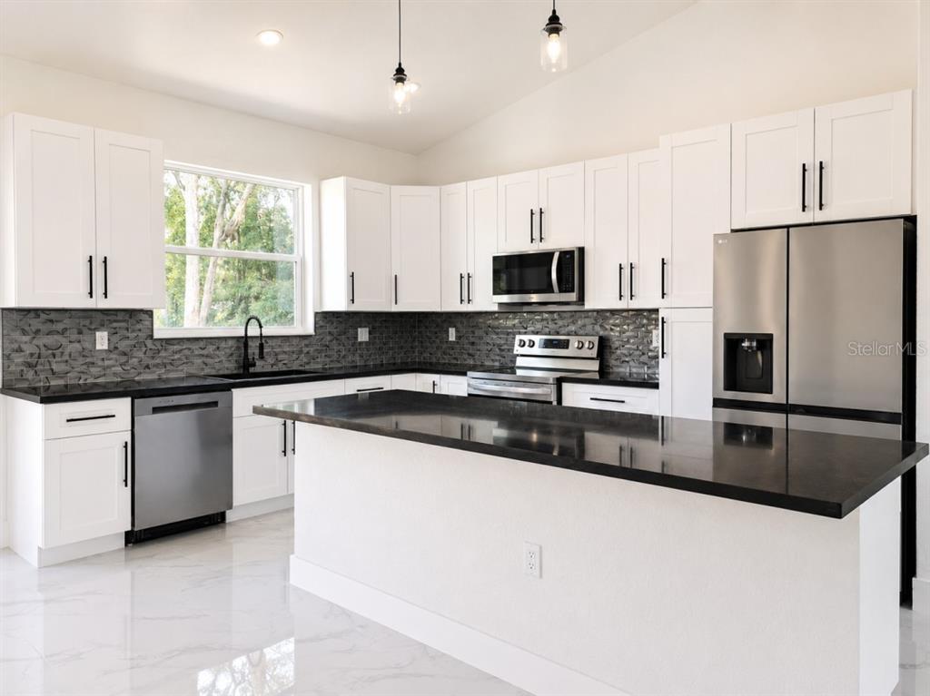 A modern kitchen features white cabinets, black countertops, stainless steel appliances, and a central island — 3476 SE HWY 484, BELLEVIEW, FL