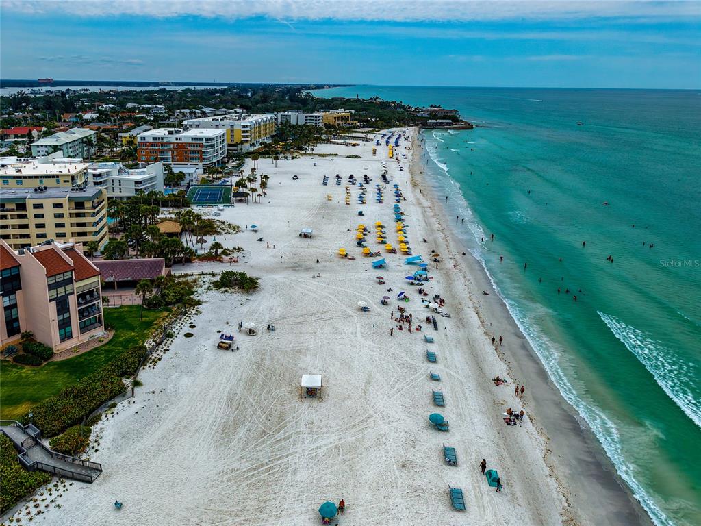 An aerial view of a sandy beach with umbrellas, lounge chairs, and nearby resort buildings — 6480 MIDNIGHT PASS ROAD UNIT 503, SARASOTA, FL