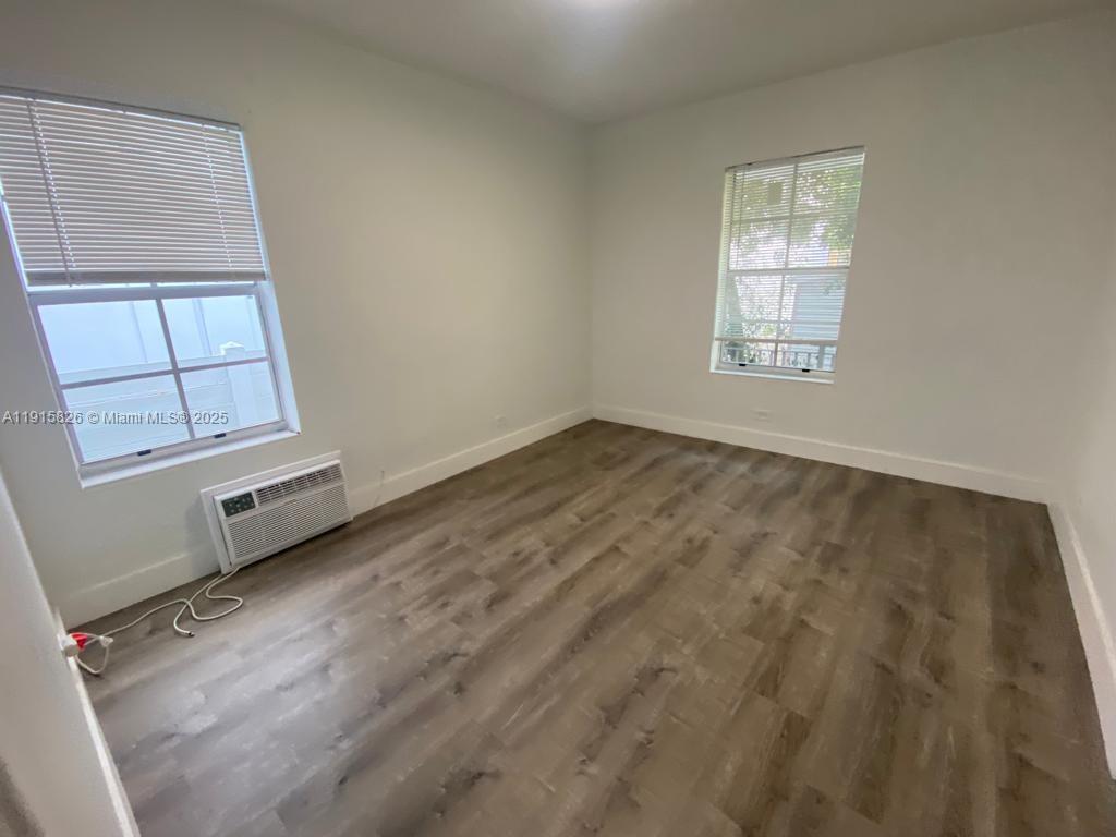 An empty bedroom features light walls, wood-look flooring, two windows with blinds, and an air conditioner — 6805 Abbott Ave # 7, Miami Beach FL 33141, MIAMI BEACH, FL